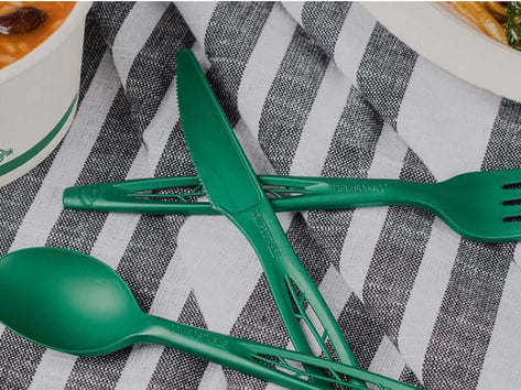 Green heavy duty cutlery set including knife, fork, and spoon on striped fabric with bowl of soup and plate of noodles in background.