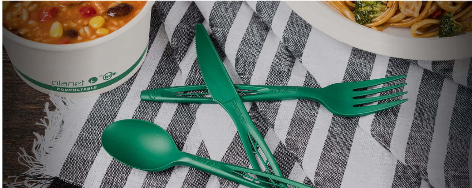 Green heavy duty cutlery set including knife, fork, and spoon on striped fabric with bowl of soup and plate of noodles in background.